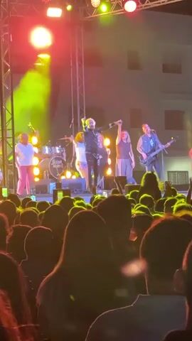 A band is performing on a stage with a crowd of people in front of them. The lead singer, a man with blond hair and a black jacket, is holding a microphone and gesturing. There are other band members on stage, including a guitarist and a drummer. The stage is lit with bright lights, and there is smoke in the air. The crowd is holding up their phones to record the performance.