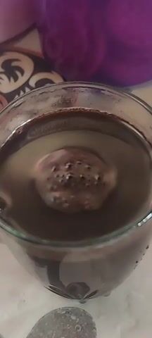 A close-up shot shows a glass of dark liquid, possibly coffee or hot chocolate, with a large ice sphere floating in the center. The ice sphere is melting, causing bubbles to form on its surface and disperse into the liquid. The glass is sitting on a table with a patterned placemat. In the background, a purple container and a white container are visible. The lighting is dim, and the focus is on the ice sphere and the drink.