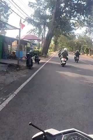 The video shows a first-person perspective of someone riding a scooter down a paved road. The rider is wearing a helmet, and the handlebars and a portion of the scooter's instrument panel are visible in the foreground. Several other scooters, each with one rider, are ahead on the road, traveling in the same direction. The road is lined with trees on both sides. To the left of the road, there appears to be a small structure with a red awning and a parked scooter. Further down the road on the left, there are utility poles and wires. The sky is bright, suggesting it is daytime. The overall scene depicts a group of people riding scooters on a rural or suburban road.