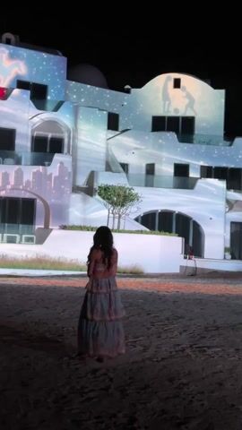 A woman with long dark hair is standing with her back to the camera in what looks like an outdoor sandy area. She is wearing a flowy layered dress. Behind her is a white building, illuminated with a light show that includes stars, clouds, and images, including what appear to be people playing. The setting is at night, and the scene is still with a voiceover playing.