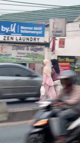 Two individuals wearing hijabs are walking across a street. The person on the left is wearing a light pink hijab and a pink and white patterned outfit. The person on the right is wearing a black hijab and a red and white striped top with white pants. Both are carrying plastic bags. They are walking in front of a row of shops. One shop is labeled "ZEN LAUNDRY" and features signs advertising "Express & Regular" laundry services. Another shop is a food stall with images of various dishes. A blue truck with "VIAR" written on it is parked in front of the food stall. A blurry motorcycle is in the foreground, suggesting the video is shot from a moving vehicle. The sky is overcast.