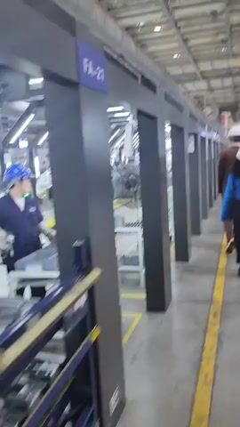 This media file shows an interior view of a car manufacturing assembly line in Yufengshan, China. The scene is well-lit with fluorescent lighting from the ceiling, suggesting it could be daytime or evening within a factory setting. Several car bodies, in a light gray primer finish, are visible on yellow elevated platforms, indicating they are in various stages of assembly. One car in the foreground is partially complete, with its doors open. A large industrial fan is positioned to the left, likely for ventilation.

There are a few workers present, one clearly visible in a blue jacket, white gloves, and a blue hard hat, actively working on or near a car body. Another figure, wearing a blue shirt, is partially visible further down the line. The atmosphere appears to be functional and industrial, with no overt emotional cues.

The setting is a modern factory with a high ceiling and exposed structural elements, typical of large-scale manufacturing facilities. The floor is marked with yellow lines, delineating pathways and work areas. There are shelves and carts with tools and parts scattered around, characteristic of an active assembly line.

Visible text includes "FA-21" and "FA-22" on blue signs mounted on the gray structural beams, which likely denote specific stations or sections of the assembly line. The overall impression is a glimpse into the organized and systematic process of automotive production.