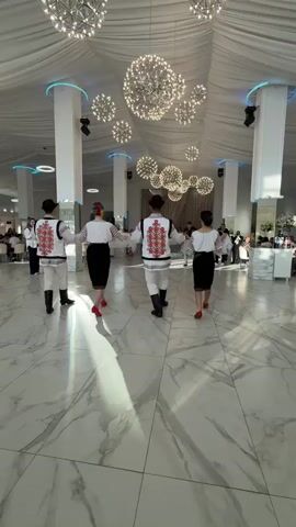 The video shows four people dancing in traditional Romanian clothing in a large, well-lit event hall. The dancers are arranged in a circle, holding hands. Two of the dancers are male, wearing white shirts with red and black embroidery, white pants, and black boots. The other two are female, wearing white blouses with embroidered sleeves and black skirts. They are also wearing colorful head coverings. The floor is made of white marble with gray veining. The ceiling is draped with white fabric and decorated with numerous spherical chandeliers that emit a warm light. Several white pillars support the ceiling. In the background, other guests are seated at tables. The lighting is bright, creating strong shadows on the floor from the dancers.