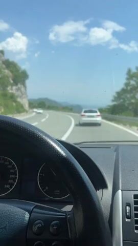 The video shows a first-person perspective from the driver's seat of a car on a mountain road. The car is following a white sedan. The scenery includes rocky cliffs, mountains in the distance, green trees, and a blue sky with white clouds.  The car's dashboard is visible, including the steering wheel and instrument panel. Music is audible in the background.