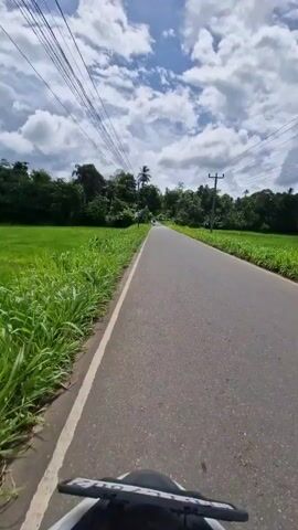 This is a first-person perspective video captured from a moving motorcycle. The video shows a paved road with a white line on the left side, bordered by lush green fields of grass on both sides. In the distance, there are trees and a few utility poles with overhead wires. The sky is blue with scattered white clouds. The motorcycle's handlebars and a portion of its front are visible at the bottom of the frame. The scene appears to be a rural landscape under a bright, partly cloudy sky.