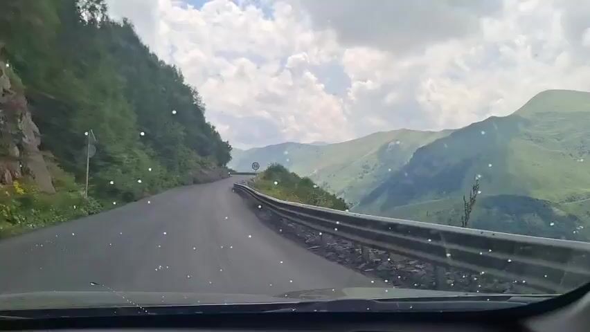 The video shows a point-of-view shot from a car driving on a winding mountain road. The road is paved and appears to be in good condition. There is a guardrail on the right side of the road, and a steep, forested mountain slope on the left. In the distance, there are rolling green mountains under a cloudy sky. A speed limit sign indicating 50 is visible. Another vehicle is seen driving ahead on the road. The windshield of the car has several small, white specks, possibly water droplets or debris. The overall scene depicts a scenic drive through a mountainous region.