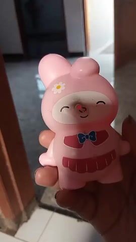A hand is holding a pink toy bunny. The bunny has a white flower with a yellow center on its forehead, closed eyes with eyelashes, a pink nose, and a blue bow tie with a white center. The bunny is wearing a pink dress with a ruffled hem. The background is out of focus but appears to be an indoor setting with a tiled floor.