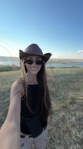 A woman in a cowboy hat and sunglasses takes a selfie while standing on a grassy hill. She is wearing a black tank top and light-colored pants. In the background, a wide river or lake stretches across the landscape under a clear blue sky. The sun is shining brightly. The woman smiles and turns her head slightly as she records the video.