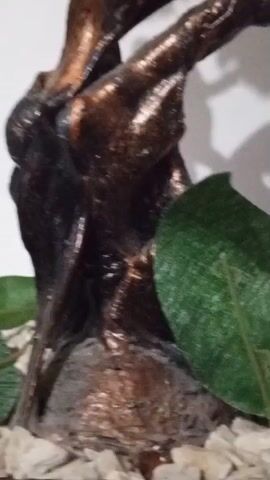 A close-up video shows a decorative object. The object appears to be a small tree sculpture with a dark, textured trunk that has a metallic bronze sheen. The trunk is twisted and has multiple branches. There are several white artificial flowers with pink centers attached to the branches. Some green artificial leaves are also visible near the base of the trunk. The base of the sculpture is decorated with small white pebbles. The background is a plain white wall, with shadows cast by the sculpture and flowers. The lighting is bright, creating highlights on the metallic trunk and the petals of the flowers. The video is shot from a low angle, looking up at the sculpture.