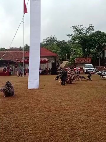 The video opens with a wide shot of an outdoor event. In the foreground, the lower half of a person's face is visible, facing away from the camera. The middle ground features a large grassy field, a tall white banner attached to a pole, and several individuals in costumes. To the left, a person in a camouflage costume, resembling a ghillie suit, is crouched low to the ground and holding a rifle. Further to the right, another individual is in a similar costume, also appearing to be in a dynamic pose. In the background, a group of people dressed in red and white outfits are gathered near a structure adorned with red and white flags. One person in this group holds a blue umbrella decorated with colorful streamers. A white vehicle is parked behind them, and other people are visible in the distance, seated and standing. The environment appears to be a rural area, with houses and trees in the background, under an overcast sky. The video then transitions to a series of close-up shots focusing on different aspects of the event. The second clip shows the same grassy field with the banner and costumed individuals. The person in the camouflage suit on the left is now standing and appears to be swinging a staff or stick. Another costumed figure is visible further back in the center. The group in red and white is still present, and the person with the umbrella is waving. The third clip zooms in on the costumed individuals in the middle ground. The person in the ghillie suit on the left is still visible, and another figure in a dark costume is in motion. The group in red and white is more prominent, and the individual with the umbrella is now closer to the camera, showcasing the details of the umbrella and streamers. The fourth clip continues to focus on the costumed performers and the group in red and white. The individuals in ghillie suits are no longer the primary focus. The group in red and white, particularly the person with the umbrella, is well-lit and clearly visible. The background shows the white vehicle and the surrounding structures. The fifth and final clip shows the same scene, with the costumed performers and the group in red and white. The person with the umbrella is still visible, as are other members of the group. The overall