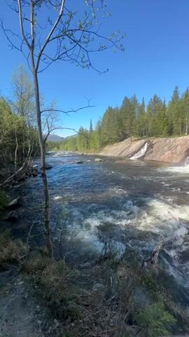 The video shows a scenic view of a waterfall flowing over rocks in a forest on a clear, sunny day. The camera pans from left to right, revealing a woman sitting on a rock near the edge of the river. She is wearing casual clothes and a face mask and waves her hand towards the camera. The surrounding environment includes trees, bushes, and a clear blue sky. The focus remains on the natural beauty of the waterfall and the woman's presence within the landscape.