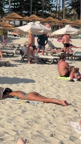 The video captures a vibrant, sunny day at a sandy beach, likely in Ayia Napa, Cyprus. The scene is bustling with numerous people enjoying their holiday.

In the initial frames, many individuals are seen relaxing on the sand. Some are sunbathing directly on towels, including a woman in a light-colored bikini lying face down in the foreground. Others are on lounge chairs under beach umbrellas. A woman in a black bikini stands in the middle ground, near a man in blue shorts. Palm trees and multi-story hotel buildings line the background.

The video then transitions to the water's edge, showcasing the clear, turquoise sea. A couple, a man in swim shorts and a white baseball cap, and a woman in a black bikini top and shorts, walk hand-in-hand along the shallow water, carrying beach items. Several other people are seen swimming, wading, or standing in the inviting sea. A prominent white hotel building, clearly displaying the text "NissiBlu" on its side, dominates the background, surrounded by more palm trees.

Further along the shoreline, more beachgoers are visible, including a woman in a light-colored top and shorts carrying a bag, walking alongside a child. The sky is bright blue and largely clear, indicating excellent weather conditions. The overall atmosphere is one of relaxation and enjoyment, with people engaging in typical beach activities like sunbathing, swimming, and strolling along the shore. The strong sunlight and visible shadows suggest it is daytime, likely mid-day or afternoon.