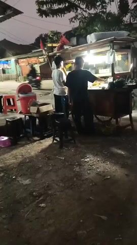 The video shows an outdoor scene at dusk or dawn. In the foreground, the ground is dirt and gravel, with some shadows cast by overhead structures. To the left, there are several plastic stools of different colors, including red and pink, and a red bucket on a small table. A box is also visible on the table. Two people are standing near a food stall, which is illuminated by overhead lights. One person, wearing a white shirt, faces the stall. The other person, wearing a dark shirt and a head covering, is behind the stall, appearing to be preparing food or serving customers. The food stall has a counter with items displayed on it. In the background, there are more buildings and trees. A motorcycle is seen passing by on the road in the distance. A pole with red and white stripes is also visible. The overall impression is that of a street food vendor operating in a modest, informal setting.