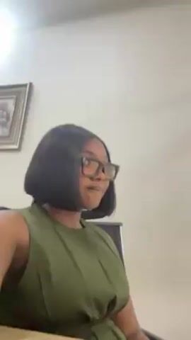 A woman with short black hair and glasses sits in an office setting. She wears a sleeveless green dress. She touches her hair with her right hand, and a watch is visible on her wrist. A framed picture hangs on the wall behind her. The lighting appears to be from overhead.