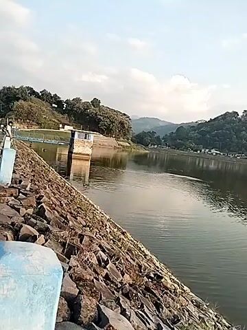 The video displays a serene landscape featuring a large body of water, likely a reservoir or dam, set against a backdrop of hills and mountains. In the foreground, a rugged, rocky embankment slopes down into the water. A paved road or path runs along the top of this embankment on the left, bordered by light blue concrete posts. A person can be seen walking in the distance on this path.

Across the water, a small, rectangular building with a blue roof and light-colored walls stands on the edge of the reservoir, connected to the land by a light blue metal bridge. The building and its reflections are clearly visible on the calm water surface. Behind this structure, the shoreline is covered with dense green trees, leading up to rolling hills and further distant mountains under a partly cloudy sky.

A dark, indistinct object, possibly an animal or debris, is seen slowly moving in the water, creating subtle ripples. The overall impression is one of peaceful natural scenery, captured during daylight hours with soft, natural lighting. No discernible text is visible within the video frame.