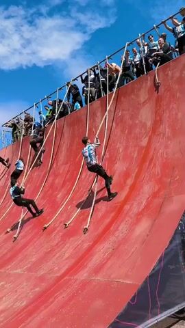 A group of people are climbing a steep, curved red wall using ropes. The wall appears to be part of an obstacle course. At the top of the wall, spectators are watching. The sky is blue with some clouds.