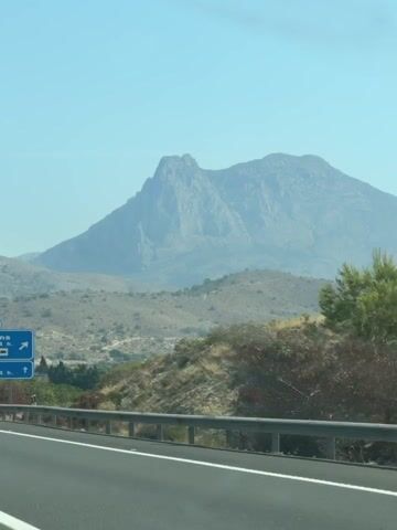The video shows a roadside view from a vehicle on a sunny day. A large mountain dominates the background, with rolling hills in the mid-ground. In the foreground, a highway guardrail runs parallel to the road. Two blue road signs are visible on the left side of the road. The upper sign indicates "La Marina" with services like petrol, parking, and restaurants, along with distances. The lower sign shows a distance of 42 km. Further down the road, another smaller sign is visible. The sky is clear and blue.