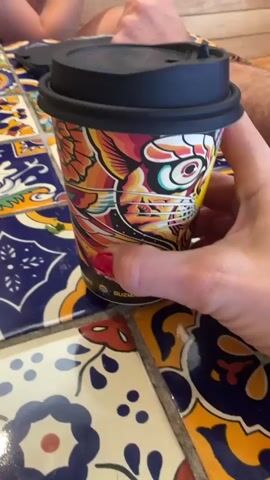The video shows a hand with red nail polish holding a coffee cup with a colorful design of a cat. The cup has a black lid and a yellow sticker with text that is partially visible. The cup is placed on a table with a tiled surface featuring blue, white, yellow, and red patterns. In the background, parts of two people are visible, one sitting to the left and another to the right. The lighting suggests an indoor setting, possibly a cafe or restaurant. The hand is rotating the cup, revealing different parts of its design and the sticker.