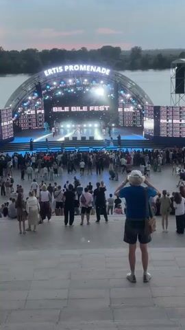 This media file captures an outdoor evening event at the Ertis Promenade in Pavlodar, Kazakhstan. The scene is dominated by a large, illuminated stage structure set against a backdrop of the Ertis River and a dusky sky. The stage features the text "ERTIS PROMENADE" prominently arched across its top, with the words "BILE BILE FEST" displayed on large screens flanking the main performance area.

A significant crowd of people is gathered on the promenade, facing the stage. They are a diverse group, dressed in casual summer attire, suggesting a warm evening. The audience is spread across various levels, with some standing closer to the stage and others further back, seated or standing on elevated platforms and staircases. In the foreground, a person wearing a white cap and blue t-shirt stands with their back to the camera, appearing to be observing the event.

The event itself seems to be a performance or concert, given the stage setup, lighting, and the assembled audience. The lighting on the stage is bright and dynamic, with spotlights illuminating performers on stage. The overall atmosphere appears festive and energetic, with the large gathering of people indicating a popular event. The time of day is evident from the twilight sky, with the transition from day to night visible. The weather appears calm, suitable for an outdoor gathering.