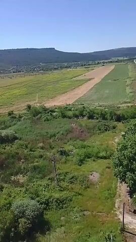 The video begins with an aerial view of a rural landscape on a clear, sunny day. Rolling green hills stretch towards the horizon, with patches of farmland and vegetation. A straight, paved road cuts through the scene, bordered by trees and fields. In the distance, a flat-topped hill rises against the blue sky. The camera pans to reveal more of the landscape, including what appears to be an abandoned airfield or a long, cleared strip of land. Further on, a train yard with several yellow train cars is visible next to industrial buildings. The video concludes by returning to the view of the road winding through the countryside, with a few small structures and trees dotting the area. The overall impression is of a peaceful, somewhat undeveloped rural environment.