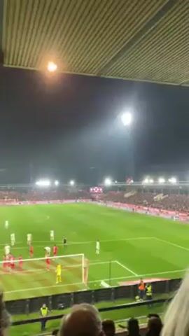 The video shows a soccer game from the stands. Players in white and red uniforms are positioned around the goal, while the goalkeeper is in yellow. The field is brightly lit, and the stands are packed with spectators. The camera is angled upwards, showing the stadium's roof and lights. The crowd is cheering loudly.