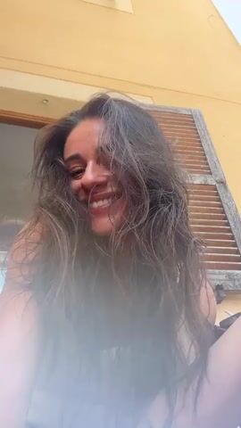 A young woman with long, dark, wavy hair is looking up at the camera and smiling. She is wearing a white tank top and a bracelet on her left wrist. Her hair is partially obscuring her face, and she is running her hand through it. The background appears to be outdoors, with a yellow wall and wooden shutters visible. There is a slight blur or lens flare effect in the foreground.