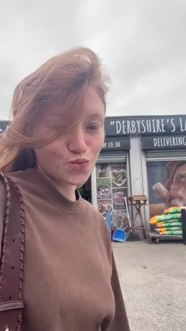 This is a selfie-style video clip featuring a young woman with auburn hair and freckles. She is wearing a brown, ribbed long-sleeved shirt and a brown bag with a braided strap is slung over her left shoulder. She is looking directly at the camera with her lips pursed.

The setting appears to be outdoors in front of a commercial building. Visible signs indicate businesses named "DERBY" and "LAVENDER DOG SHOP". There are vehicles in the background, including a white SUV and a dark blue van, suggesting a public area like a street or parking lot. The sky is overcast, indicating a cloudy day. The overall atmosphere seems casual and everyday.

The woman's expression is neutral to slightly playful. The clip is brief, showing minimal movement and activity beyond the slight sway of her hair in the breeze. The time of day is indeterminate but likely daytime due to the natural light. The visible text provides a strong location cue for Amber Valley, United Kingdom.