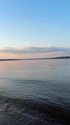 This is a vertical, first-person view of a tranquil body of water, likely a lake or river, at either sunrise or sunset. The sky is a clear, pale blue with soft, wispy pink and white clouds stretched horizontally across the upper portion of the frame. The water in the foreground is dark and rippling, with small waves lapping gently at the unseen shore. Further out, the water reflects the pastel hues of the sky, creating a shimmering, pinkish-orange surface. The horizon line is low and distant, marked by a dark, treeline silhouette on the far bank. There are no people or man-made objects visible in the frame. The overall impression is one of peace and natural beauty.