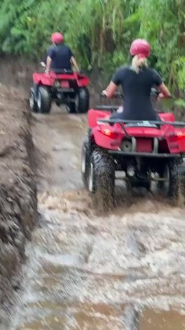 Two people on red ATVs are riding through a muddy, water-filled trail. They are wearing red helmets. The person in front is wearing a dark shirt and the person behind is wearing a black shirt. The trail is surrounded by lush green vegetation. The ATVs are kicking up water and mud as they move.