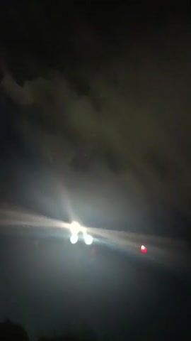 This video shows an airplane flying at night. The camera is looking up at the airplane as it passes overhead. The airplane is illuminated by its own lights and lights from the ground. The sky is dark and there are some clouds visible. The airplane appears to be descending or landing.