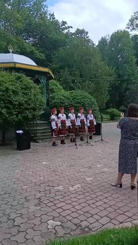 A group of five young girls wearing traditional embroidered clothing, possibly Ukrainian, stands in a line in front of microphones on a paved outdoor area. They appear to be singing. To the right of the girls, a woman in a patterned dress is taking a picture or video of them with her phone. Behind the girls is a bandstand covered in greenery, and a large number of trees form the backdrop. The sky is overcast. The ground is a decorative brick or stone pattern. There are speakers to the left and right of the performers.