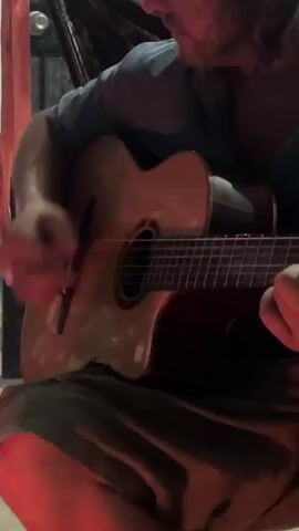 A man is sitting and playing an acoustic guitar. He is wearing a light blue t-shirt and dark shorts. The guitar is a light brown color. There are some items hanging in the background including a hammock. A red light is shining on the scene.