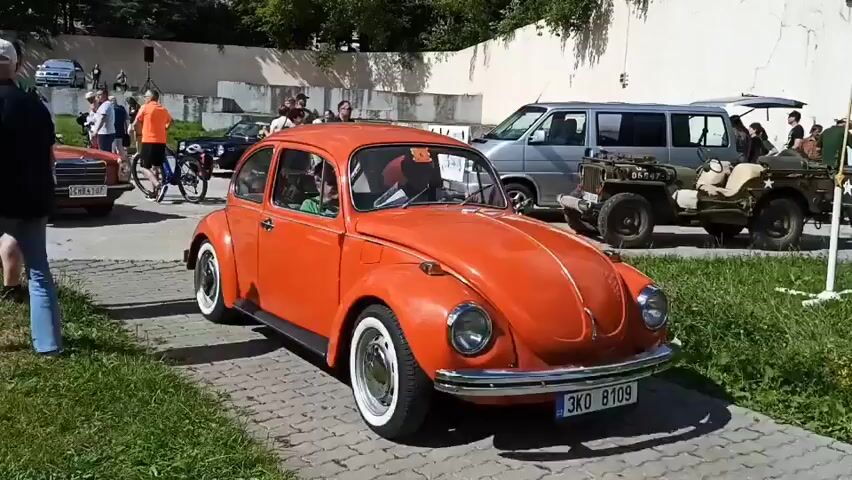 The video shows an outdoor car show. In the foreground, a bright orange Volkswagen Beetle is parked, facing right. The car has white-wall tires and chrome accents. A person with short hair wearing glasses and a green shirt is visible driving the Beetle. To the left of the Beetle, an orange Mercedes-Benz sedan is parked, facing forward. A man wearing a green t-shirt and dark shorts stands to the left of the Mercedes. Several other people are scattered around the scene, some looking at the cars. In the background, a silver van, a military-style Jeep, and a red convertible are visible. The ground is paved with bricks, and there is grass on the left side of the frame. Trees and a wall are visible in the background. The sky is bright, suggesting daylight.