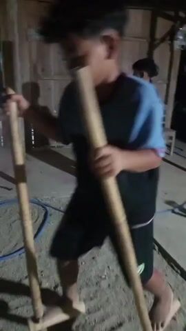 A young boy wearing a blue and black t-shirt and black shorts is standing on stilts made of bamboo. He is holding onto the stilts with his hands and his feet are resting on wooden footrests attached to the bamboo poles. The boy is in an outdoor setting with a sandy ground and some scattered debris. He appears to be walking or practicing on the stilts, as he is moving his legs and shifting his weight. The video captures him from the waist up, with the stilts extending down to the ground. The lighting is dim, suggesting it might be evening or indoors with limited light.
