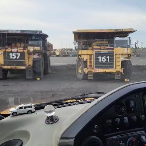 The video pans across the dashboard of a vehicle, showing a miniature white SUV and a glass sphere. The camera then moves to capture the view through the windshield, revealing a large, open-air mine or industrial site. Two massive yellow mining trucks are prominently visible, numbered 157 and 161. Other mining vehicles and structures can be seen in the background, along with a cloudy sky. The ground appears to be dirt or gravel, consistent with a mining environment.