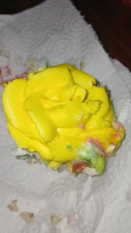 A close-up shot shows a cupcake with bright yellow frosting piped in a rose shape, sitting on a white paper towel. Crumbs and some small specks of red, green, and white are visible around the base of the cupcake. The countertop underneath is dark, out of focus. The lighting is slightly dim. The camera zooms in slowly on the cupcake.