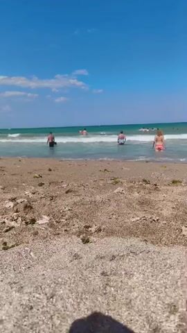 The video shows a beach scene on a sunny day. The foreground is dominated by the sand and small pebbles of the beach. In the middle ground, several people are visible in the shallow water, wading and swimming. Waves are breaking gently on the shore. In the background, the ocean stretches out towards the horizon under a clear blue sky with a few scattered white clouds. The overall atmosphere is one of leisure and recreation.