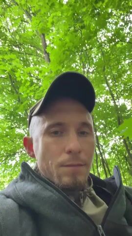 A man wearing a dark baseball cap and a grey jacket is in a forest. He is looking directly at the camera and holding a metallic cup to his lips, drinking from it. He takes a drink and then pulls the cup away from his mouth, looking back at the camera with a slight smile. The background is filled with lush green trees and foliage, suggesting a natural, outdoor setting.
