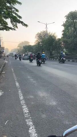 The video shows a view from the side of a road, looking forward at a line of motorcycles and cars moving in the same direction. The road is paved and has a white line marking the edge of the lane. There are trees and buildings on the left side of the road and trees on the right side. The sky is hazy, suggesting it might be early morning or late afternoon. The motorcycles are of various types, and some have passengers. One motorcycle in the foreground has a license plate that reads "W 5514 NCD". Another motorcycle further up the road has a license plate that reads "W 2457 PK". The people on the motorcycles are wearing helmets and casual clothing. The overall scene depicts a typical traffic situation in an urban or suburban area.