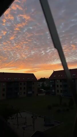 The video shows a sunset viewed through a window. The sky is filled with clouds that are illuminated by the setting sun, creating a vibrant orange and pink glow. In the foreground, a dark silhouette of a window frame obscures part of the view. Below the sky, there are two apartment buildings with red tiled roofs and dark facades. A patch of green grass lies between the buildings, and a playground structure is visible in the lower left corner. The overall scene depicts a tranquil evening in a residential area.