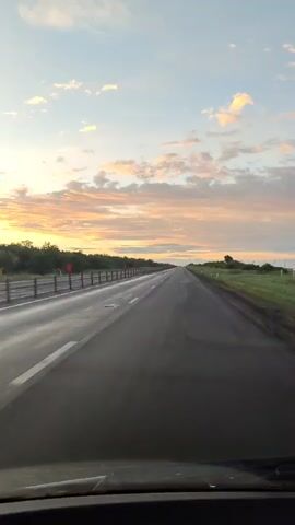 This is a first-person point-of-view video of driving on a highway at sunset. The road is paved and appears wet, reflecting the warm hues of the sky. The sky is filled with soft, scattered clouds tinged with orange and yellow from the setting sun. On the left side of the road, there is a line of orange traffic cones or markers with reflective tape, followed by a low line of vegetation. The right side of the road is bordered by green grass and some scattered trees. The video captures the vastness of the open road stretching towards the horizon under a beautiful sky.