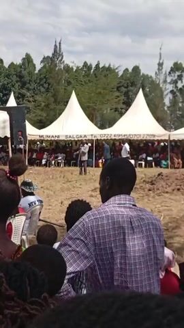 The video depicts an outdoor gathering, possibly a funeral or memorial service, with attendees seated under large white tents. A man stands in front, speaking into a microphone. The camera focuses on a person in the foreground, wearing a plaid shirt, their back to the viewer. The event appears to be taking place in a grassy field, surrounded by trees. The tent is labeled "MLIMANI SALGAA" along with a phone number. People are seated in rows of chairs, listening to the speaker. The general atmosphere seems somber and respectful.