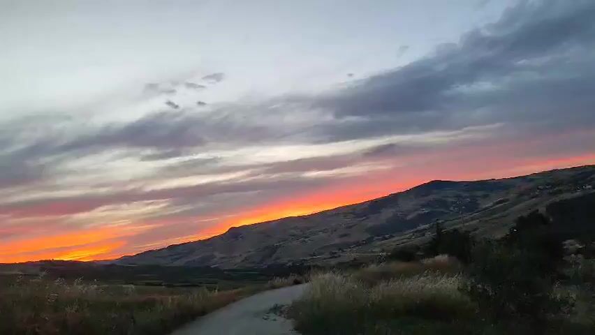 A wide shot captures a rural landscape at dusk. The sky is a dramatic spectacle of color, transitioning from soft grays and purples at the top to vibrant oranges and reds near the horizon. Wispy clouds stretch across the sky, catching the last rays of sunlight. A rolling hill dominates the right side of the frame, its slopes dotted with small clusters of trees and the faint outlines of buildings. The foreground is a dirt path, curving to the right and leading into the scene. Tall, dry grass lines the edges of the path, swaying gently in the evening breeze. The overall impression is one of natural beauty and tranquility.