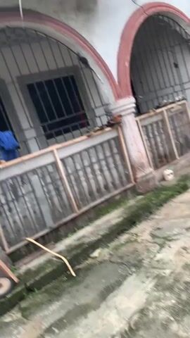 The video pans across the exterior of a building, focusing on a section with arched windows that are barred. The building appears to be aged, with some wear visible on the exterior walls and the ground in front of it is wet and appears to be a concrete patio or walkway. Various items are scattered on the ground, including what looks like cleaning supplies, discarded materials, and some gardening equipment like a wheelbarrow. There are also some clothes hanging on a railing. The overall impression is one of a somewhat neglected or lived-in outdoor space.