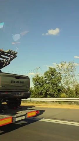 A two-level car carrier truck drives down a highway, transporting two Ram TRX pickup trucks. The truck is moving from left to right in the frame, with the camera capturing the scene from a slightly elevated position. The sky is blue with scattered clouds, and the landscape to the right of the highway consists of green fields and trees. The first Ram TRX, colored olive green with a black trim, is positioned on the lower deck of the carrier. Its rear is visible, showing the "TRX" badging on the tailgate and the distinctive taillights. The second Ram TRX, appearing dark gray or black, is on the upper deck, partially visible behind the first truck. The carrier truck itself is gray and features the "MEC" logo on its cab. The wheels of the trucks and the carrier are secured with red straps. The video appears to be filmed from another moving vehicle on the highway.