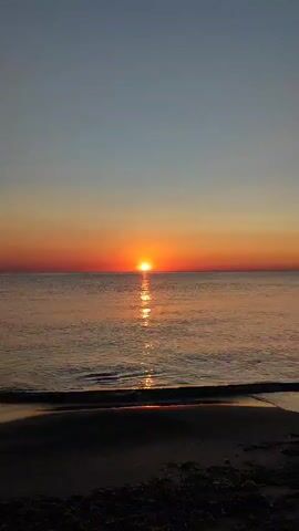 A vertical video shows a beach at sunrise. The sun is a bright orange ball just above the horizon, casting a shimmering reflection on the calm ocean. The sky is a gradient of blue and orange. Gentle waves lap the shore, which is dark and sandy.