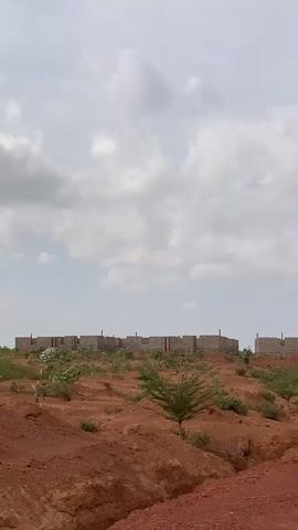 The video shows a dirt road with a reddish hue running through a sparsely vegetated landscape. In the background, there are rows of unfinished structures, appearing to be building foundations or partially constructed homes made of concrete blocks. The sky is overcast with a mix of white and gray clouds, suggesting a bright but possibly slightly cloudy day. The vegetation includes small, scattered bushes and some newly planted trees. The overall impression is of a developing area or construction site in a relatively dry environment.