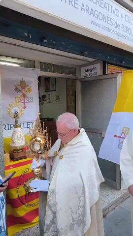 A priest in white and gold vestments holds a gold monstrance outside a building. He is wearing white gloves. Another priest in a white robe stands next to him, holding a censer. The building has a sign above its entrance. A yellow and white flag with a coat of arms is visible on the right. A red and yellow striped flag with a coat of arms is visible on the left.