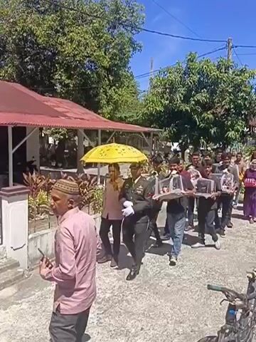 A procession is moving down a paved outdoor path. At the front of the procession is a person in a military uniform carrying a framed item. They are accompanied by another person holding a large, yellow umbrella over them. Behind them, a line of people is walking, most of them carrying framed items in clear plastic cases. The people are diverse in age and gender. The environment appears to be a sunny day with trees and buildings visible in the background. The path is bordered by a wall on the right side and a building with a red roof on the left. A bicycle is parked to the right.
