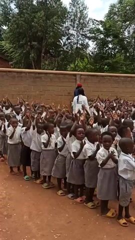 A large group of young children, presumably students at a school, are standing in a long line outdoors. They are uniformly dressed in white shirts and grey bottoms. Most of them wear sandals. An adult in a white coat and a red head covering stands amidst the children. Many of the children have their arms raised in the air. The background includes a brick wall and green trees. The ground is reddish-brown in color. At the beginning of the video, a loud sound like shouting occurs.